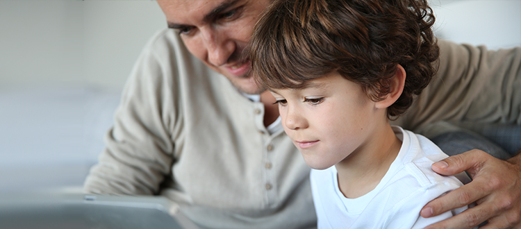 Vater und Sohn sitzen vor einem Bildschirm, während der Vater dem Kind lächelnd über die Schulter schaut. Seine Hand liegt auf der linken Schulter des Jungen. Links im Bild ist das Logo der Erich Thor Wohnungsunternehmen GmbH mit weißer Schrift auf blauem Hintergrund zu sehen.