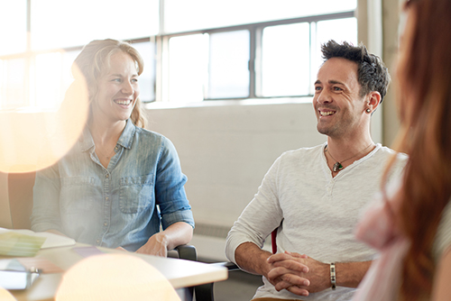 Eine Mitarbeiterin und ein Mitarbeiter sitzen in einem Meeting an einem Konferenztisch und unterhalten sich lächelnd.
